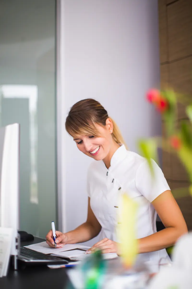 A friendly professional smiling at the modern reception desk of a Confidentures dental clinic, representing the welcoming team across their Henderson, Rosedale, and Silverdale clinical hubs.
